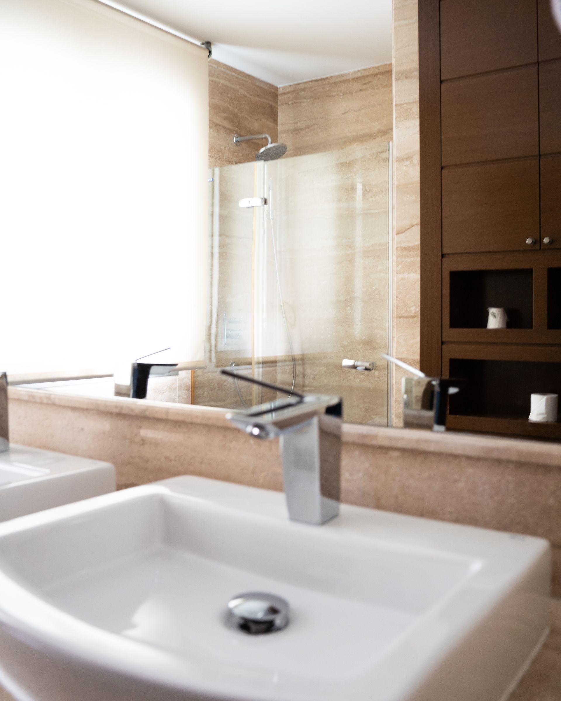 Baño con lavabos blancos, grifería cromada, paredes y ducha de mármol color canela. Mueble de madera oscura a la derecha.