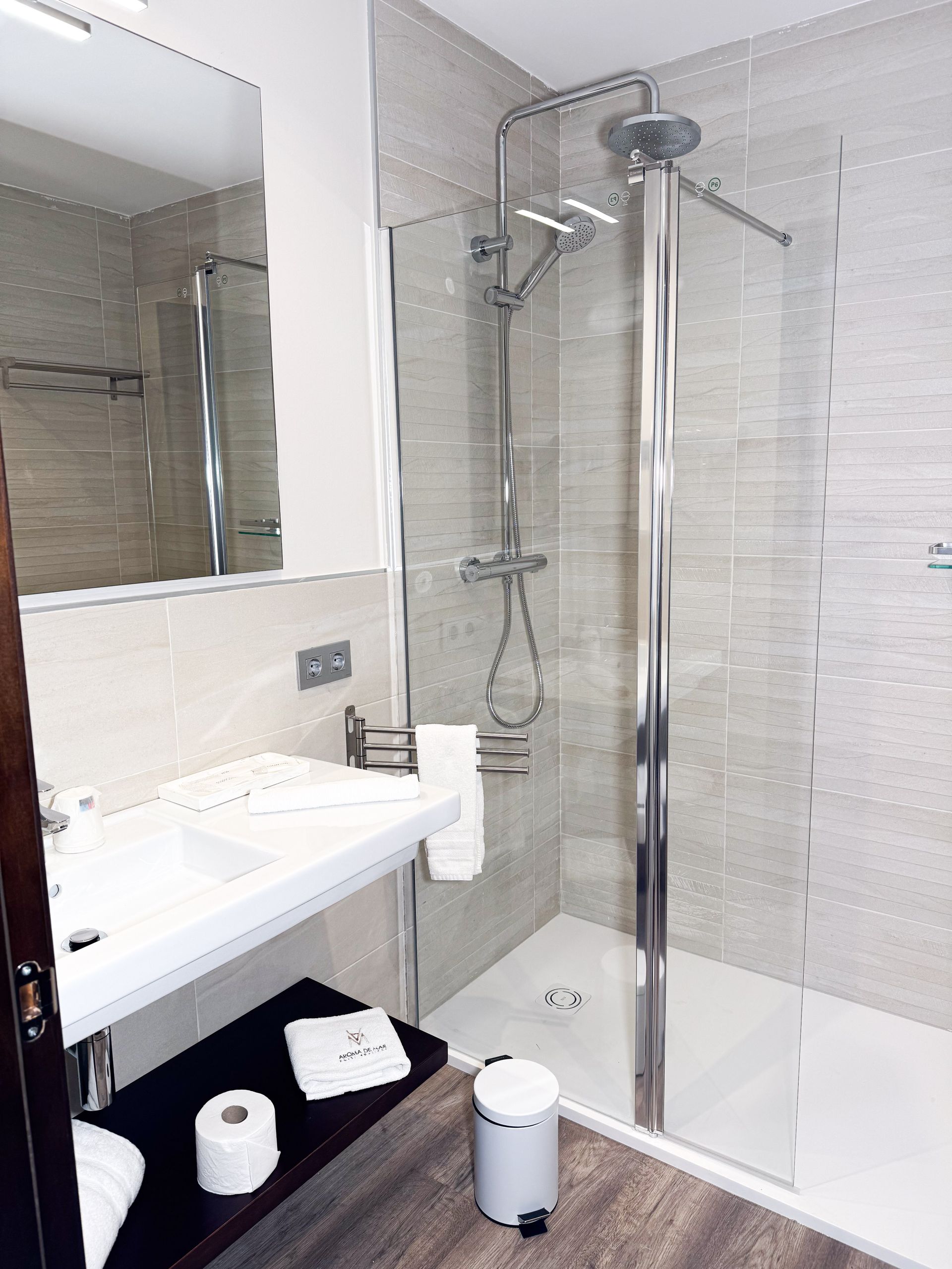 Baño con ducha de cristal, lavabo blanco y un estante oscuro con artículos de tocador.