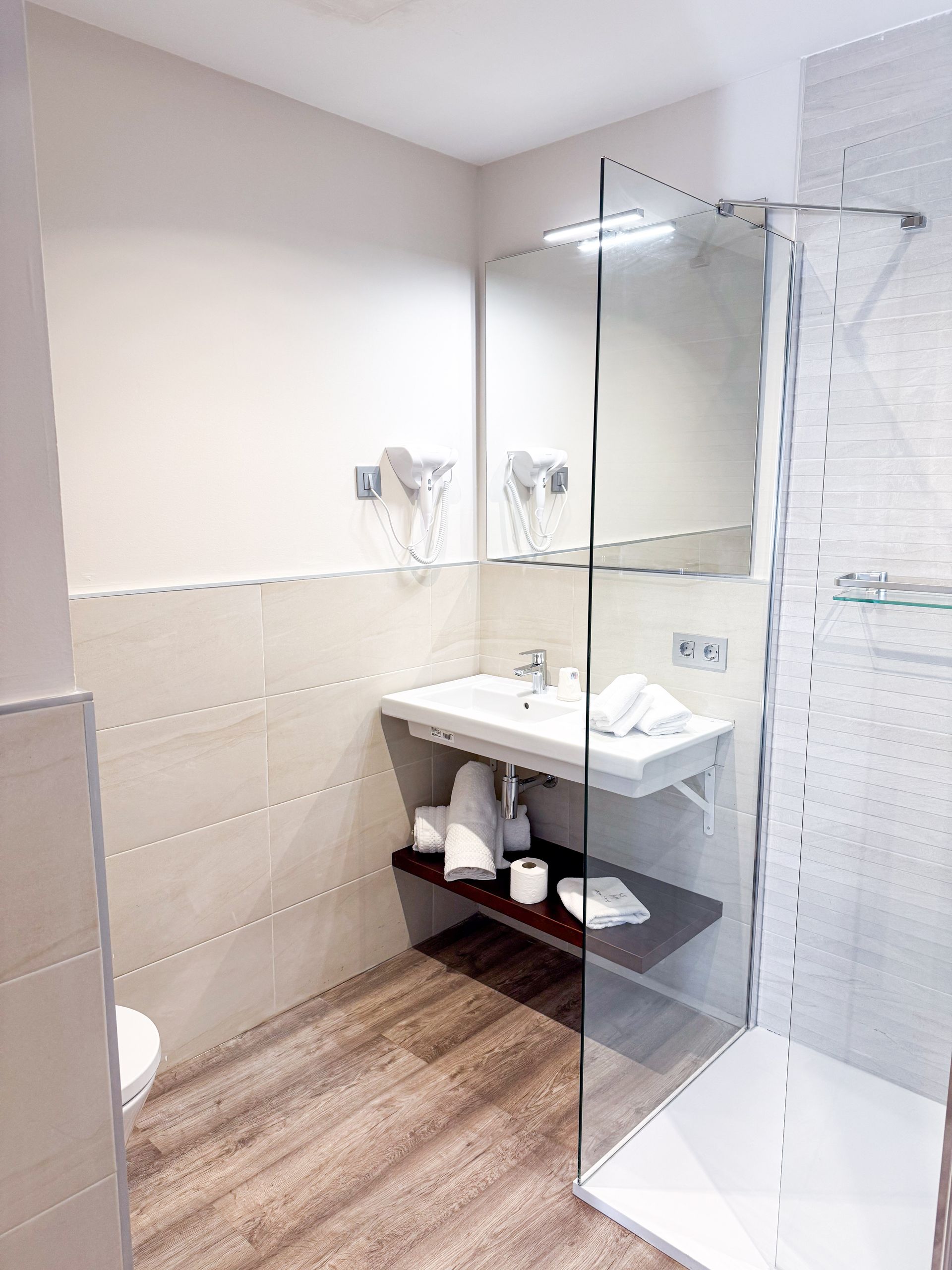 Un baño con lavabo, ducha e inodoro. Dispone de azulejos claros y mampara de cristal.