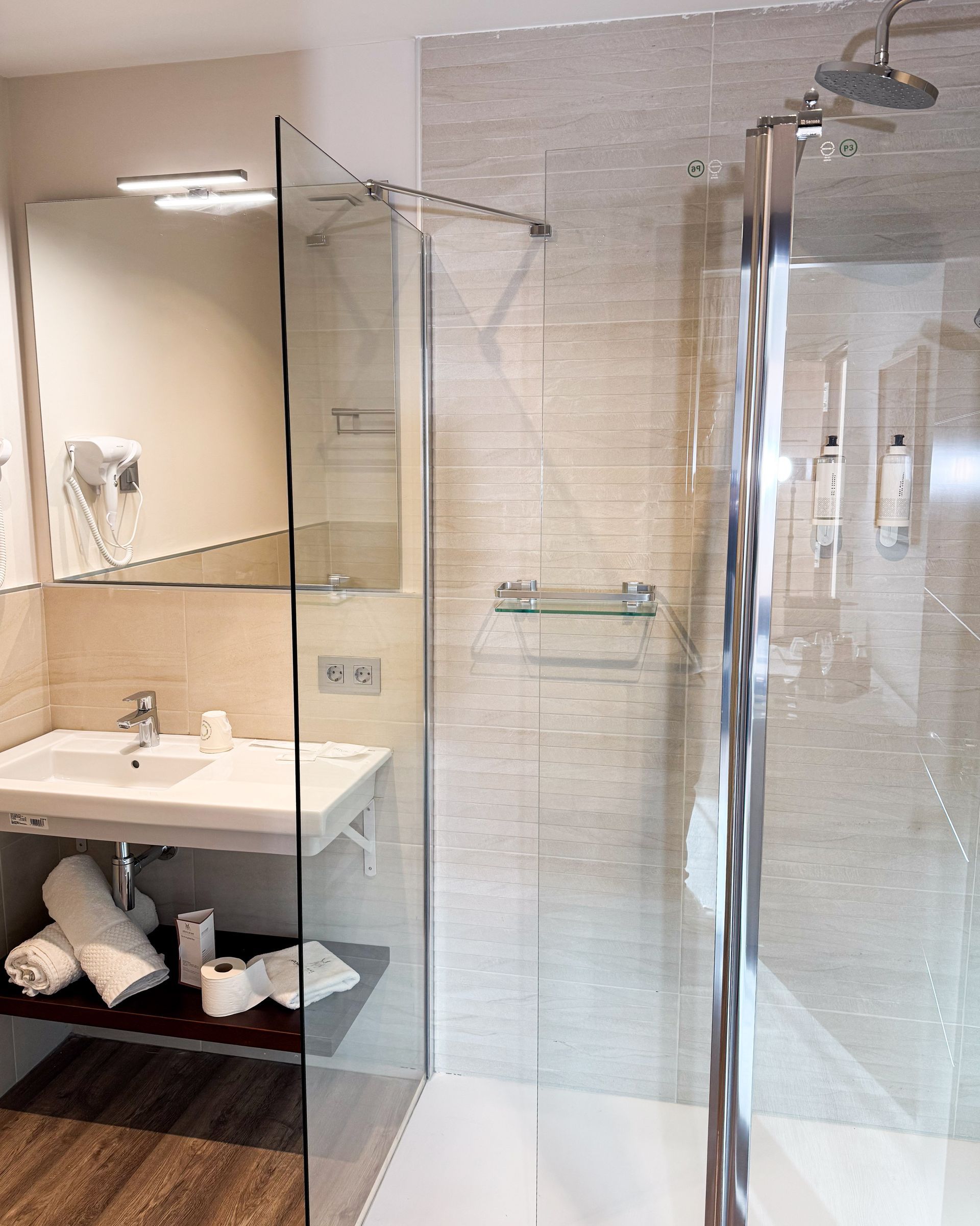 Baño con ducha de cristal, lavabo con toallas y espejo en una pared de azulejos beige.