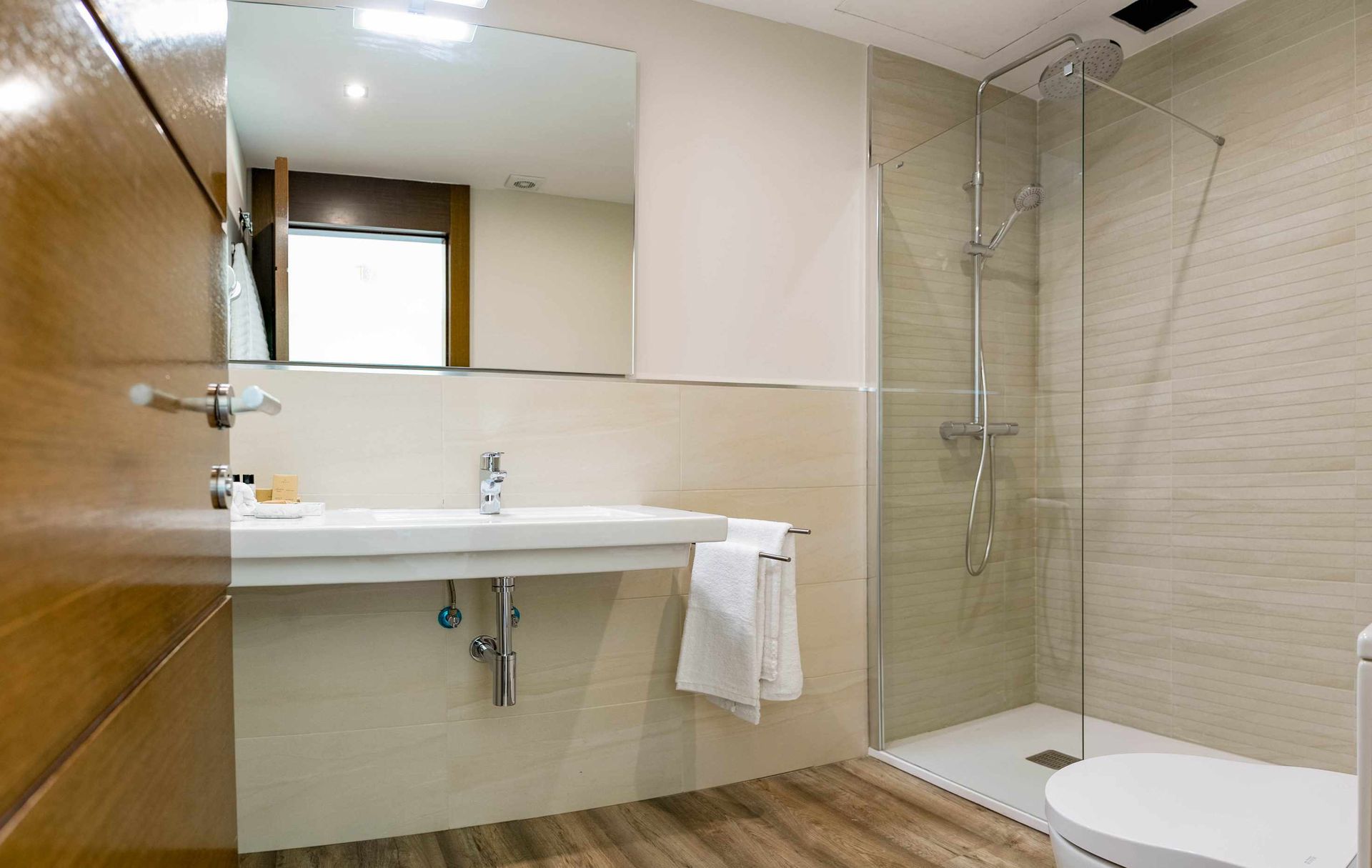 Baño moderno con lavabo, ducha e inodoro. Paredes claras, puerta de madera y un gran espejo.