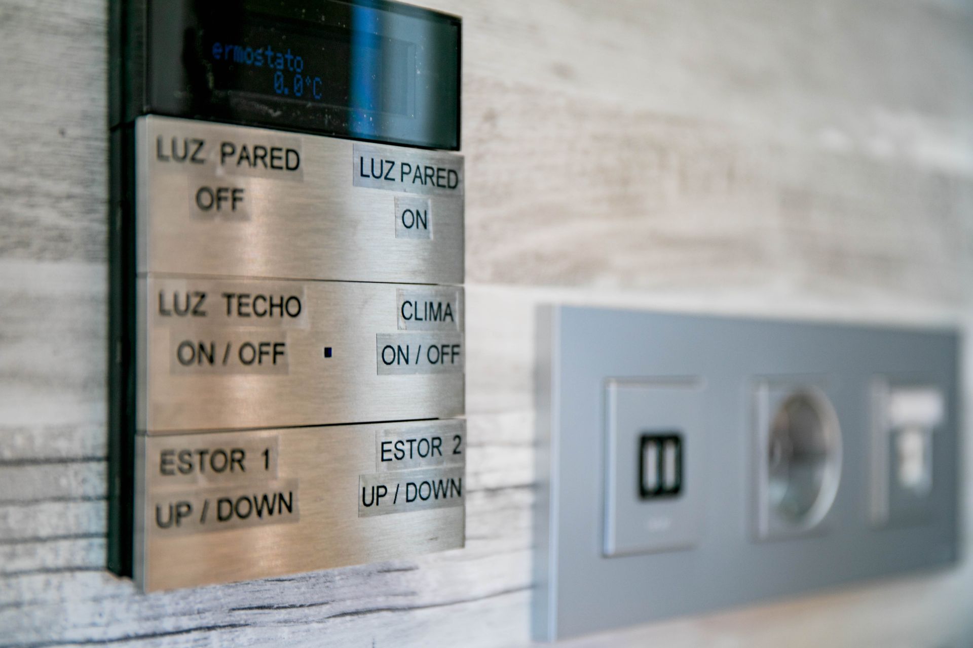 Panel de interruptores de pared plateado con botones etiquetados y una toma de corriente adyacente.