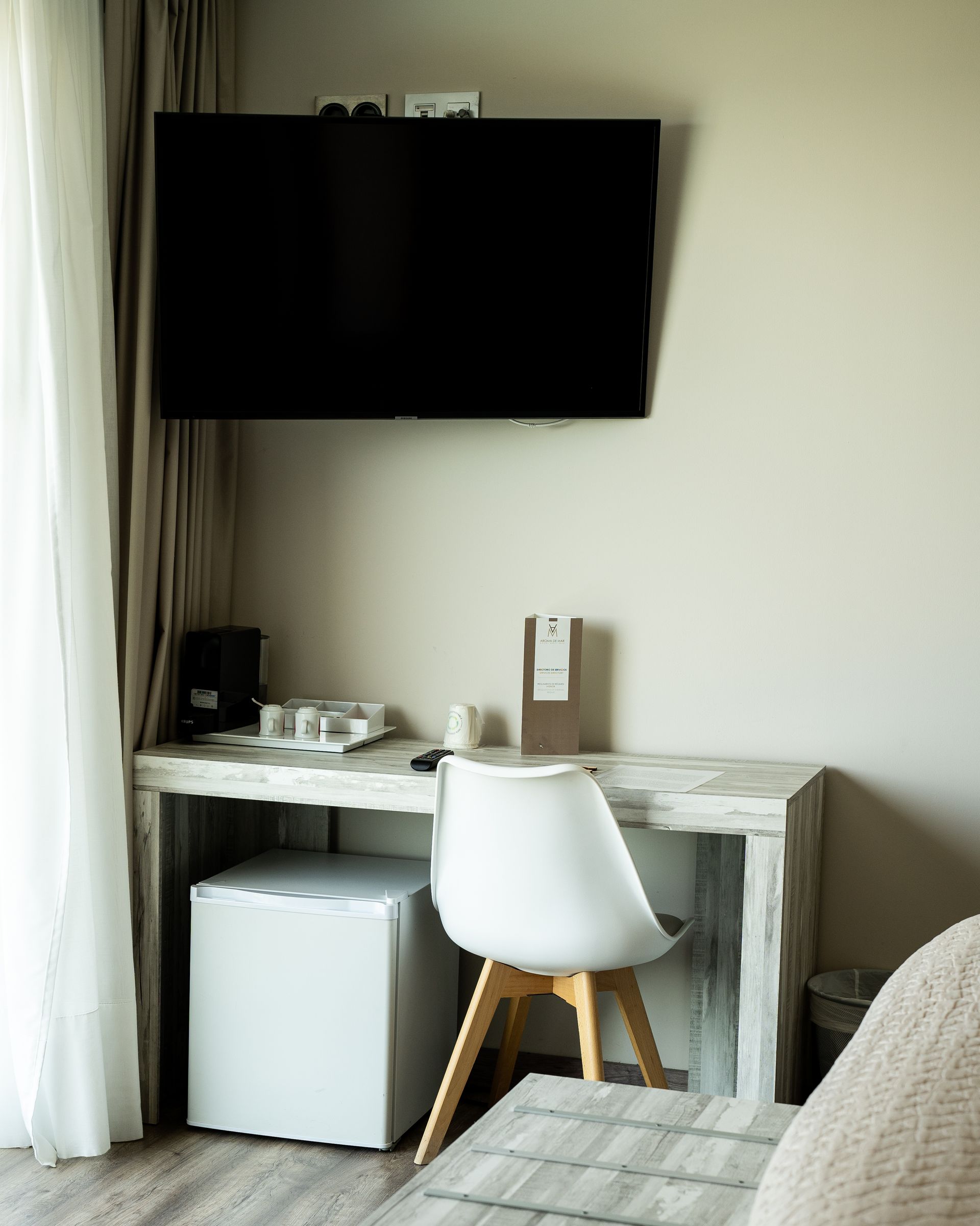Una habitación de hotel con un escritorio, un mini-nevera, una silla y un televisor en la pared cerca de una ventana.