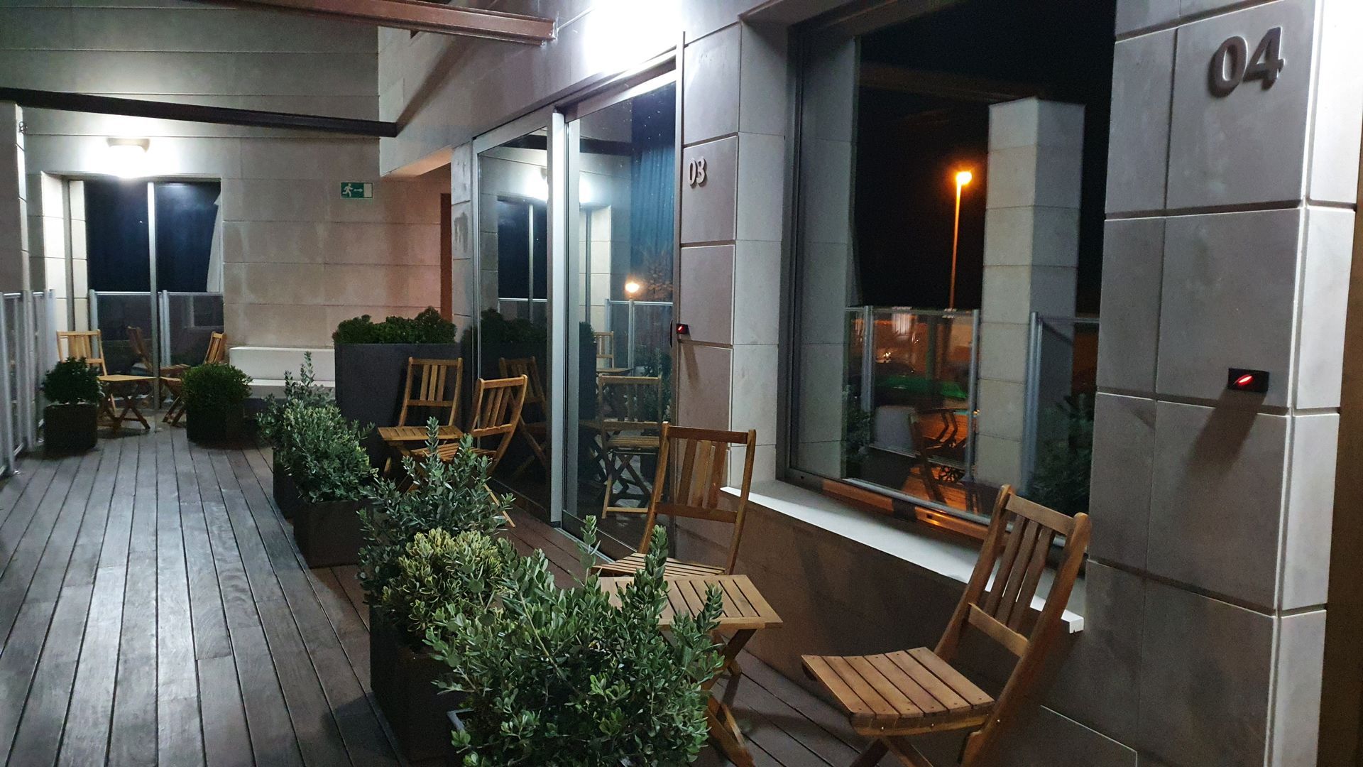 Zona de estar al aire libre con sillas de madera, vegetación y puertas abiertas por la noche.