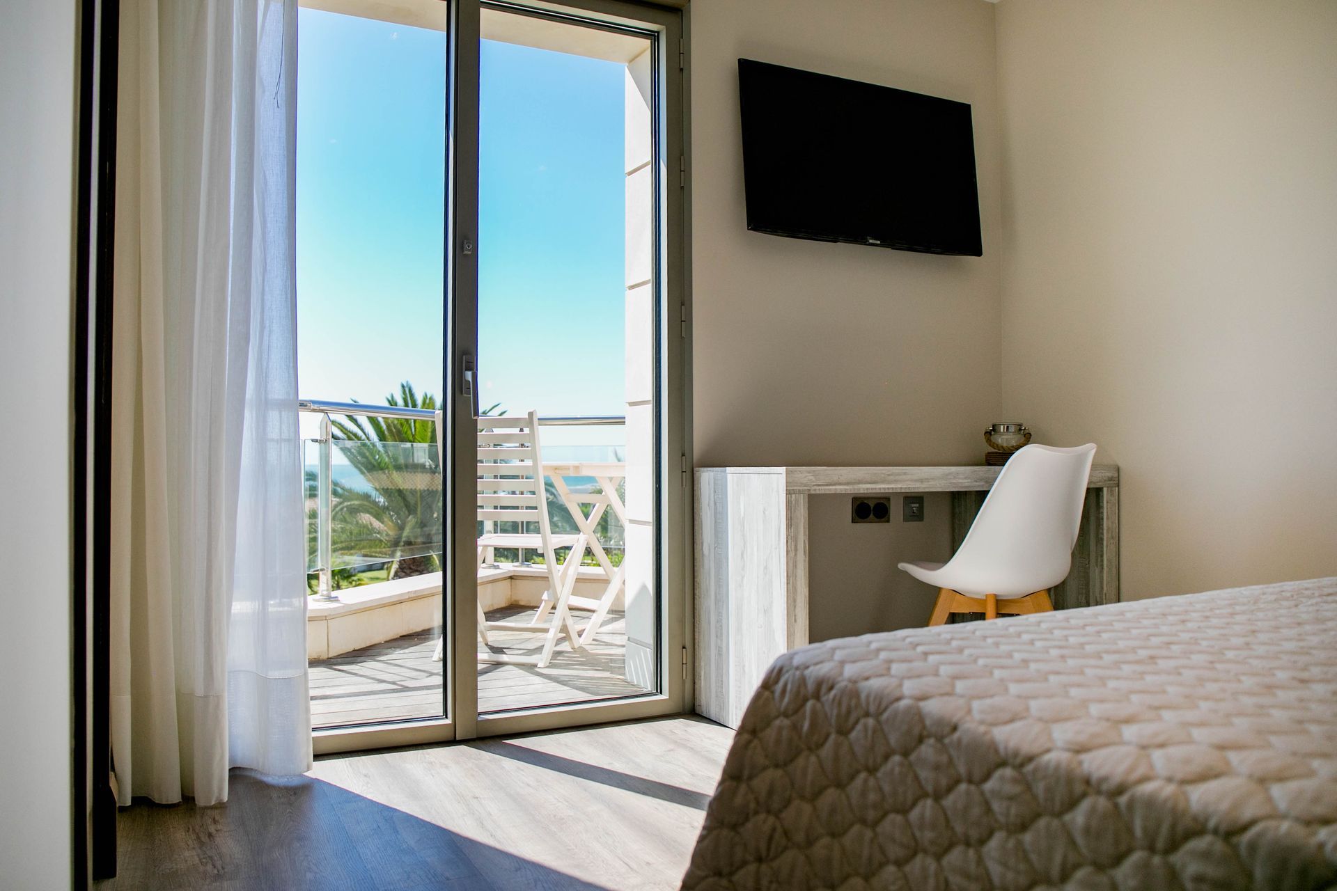Dormitorio con balcón con vista al mar. Silla blanca, escritorio pequeño, televisor y cama.
