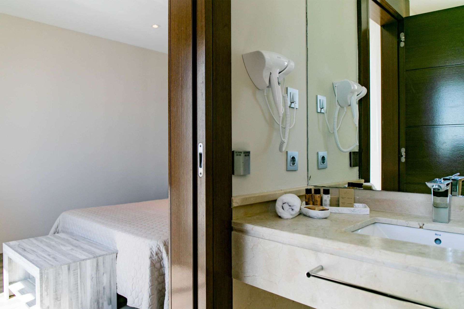 Baño con lavabo de mármol y espejo. Dormitorio visible a través de la puerta.