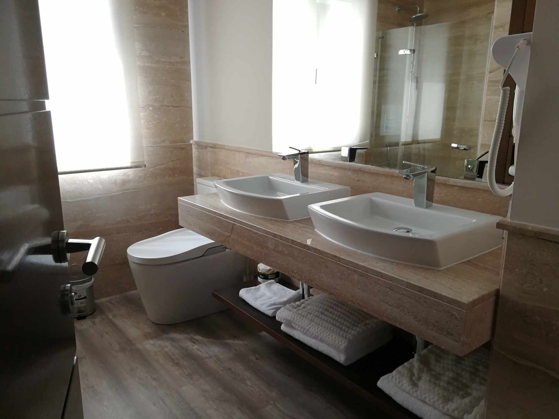 Baño con dos lavabos, inodoro y un gran espejo. Paredes color crema y suelo imitación madera.