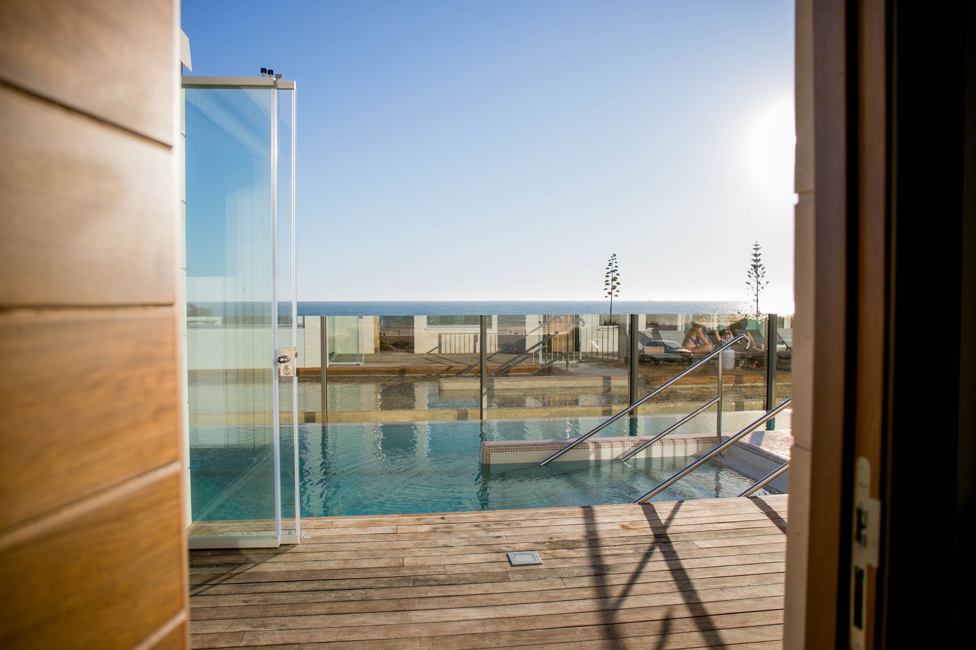 La puerta de madera se abre a una piscina con vista al océano en un día soleado.