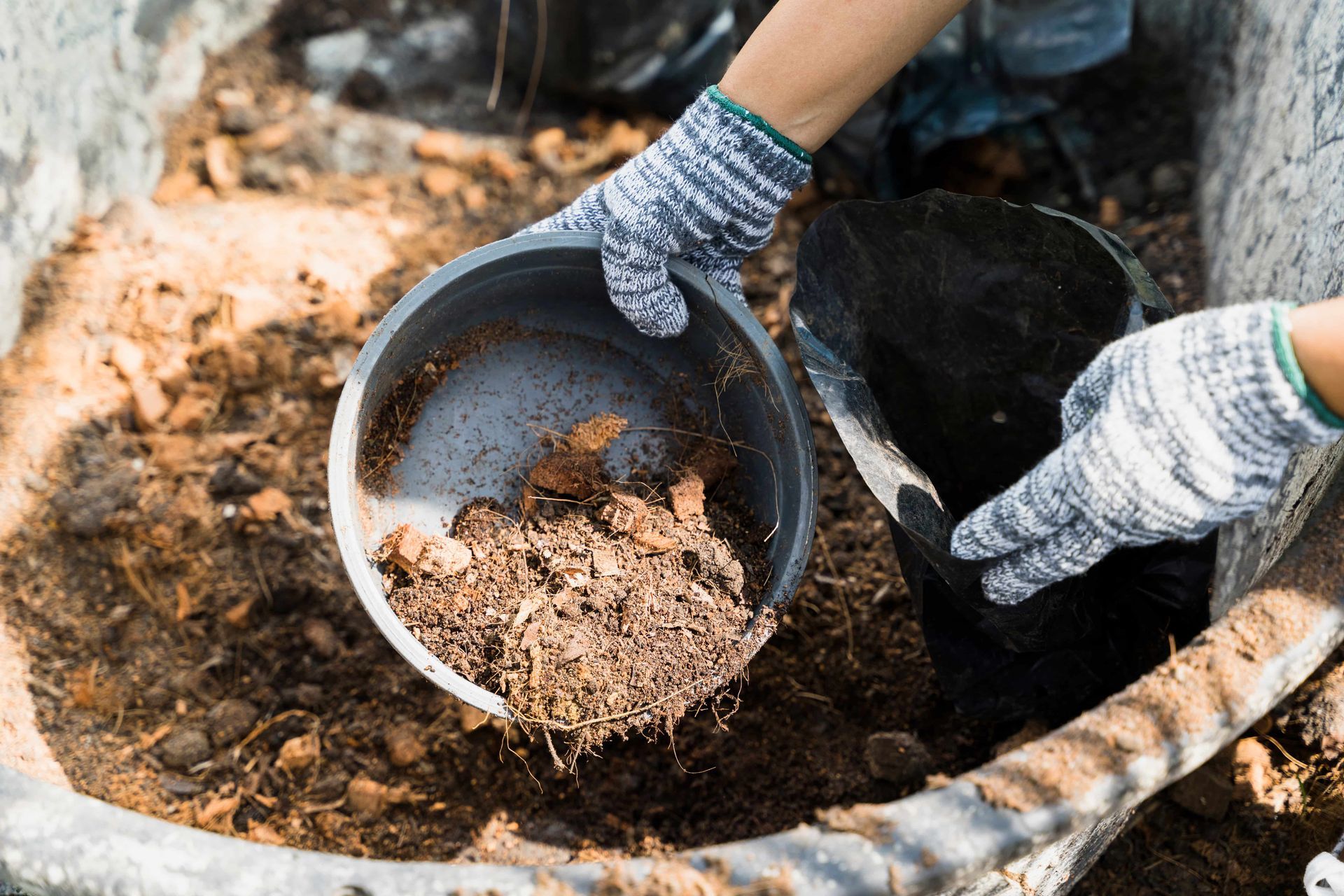 Hands wearing gloves pouring compost from a container into a larger bin.