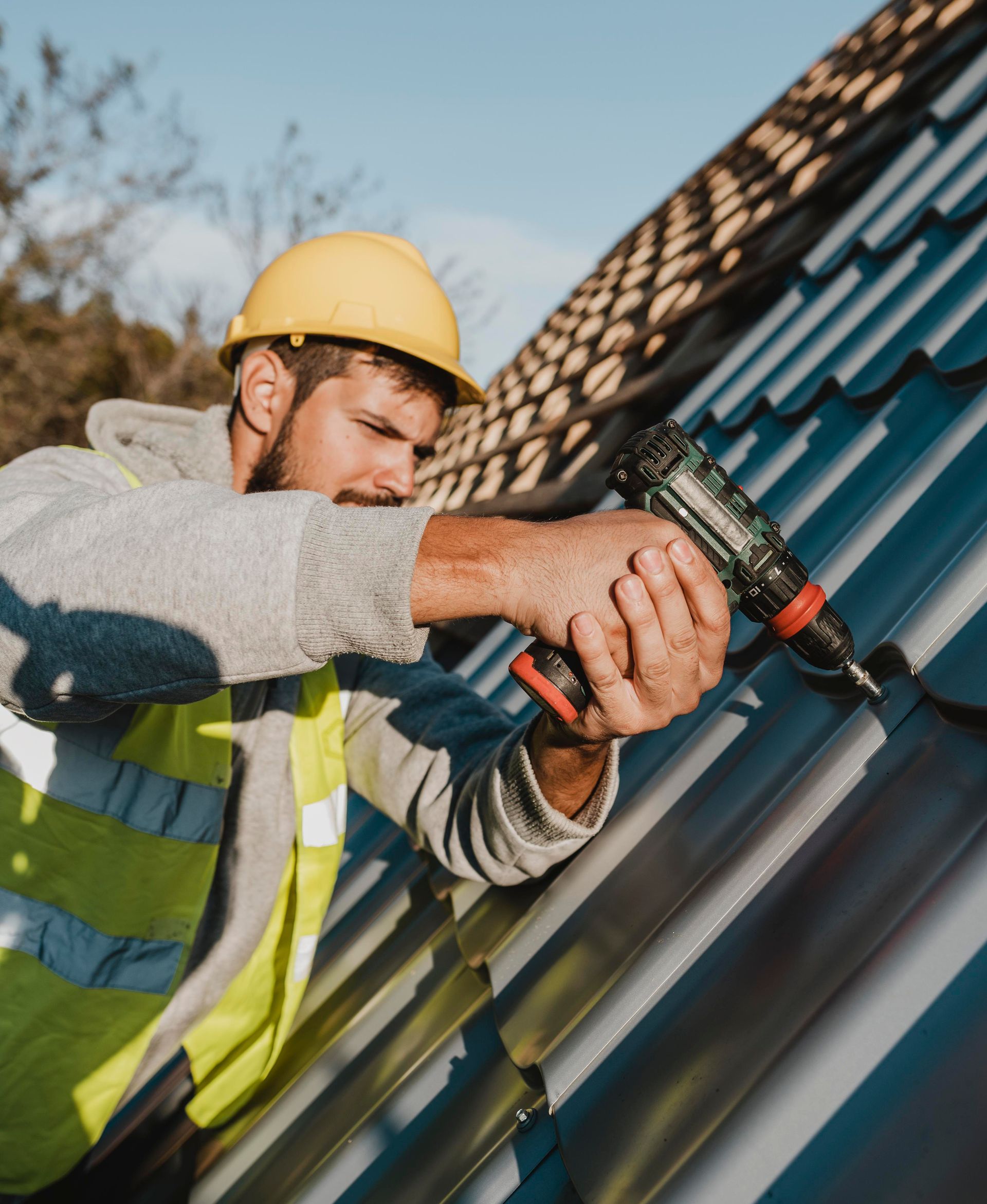 Roofer in yellow hard hat, using a drill to install metal roofing on a sunny day.