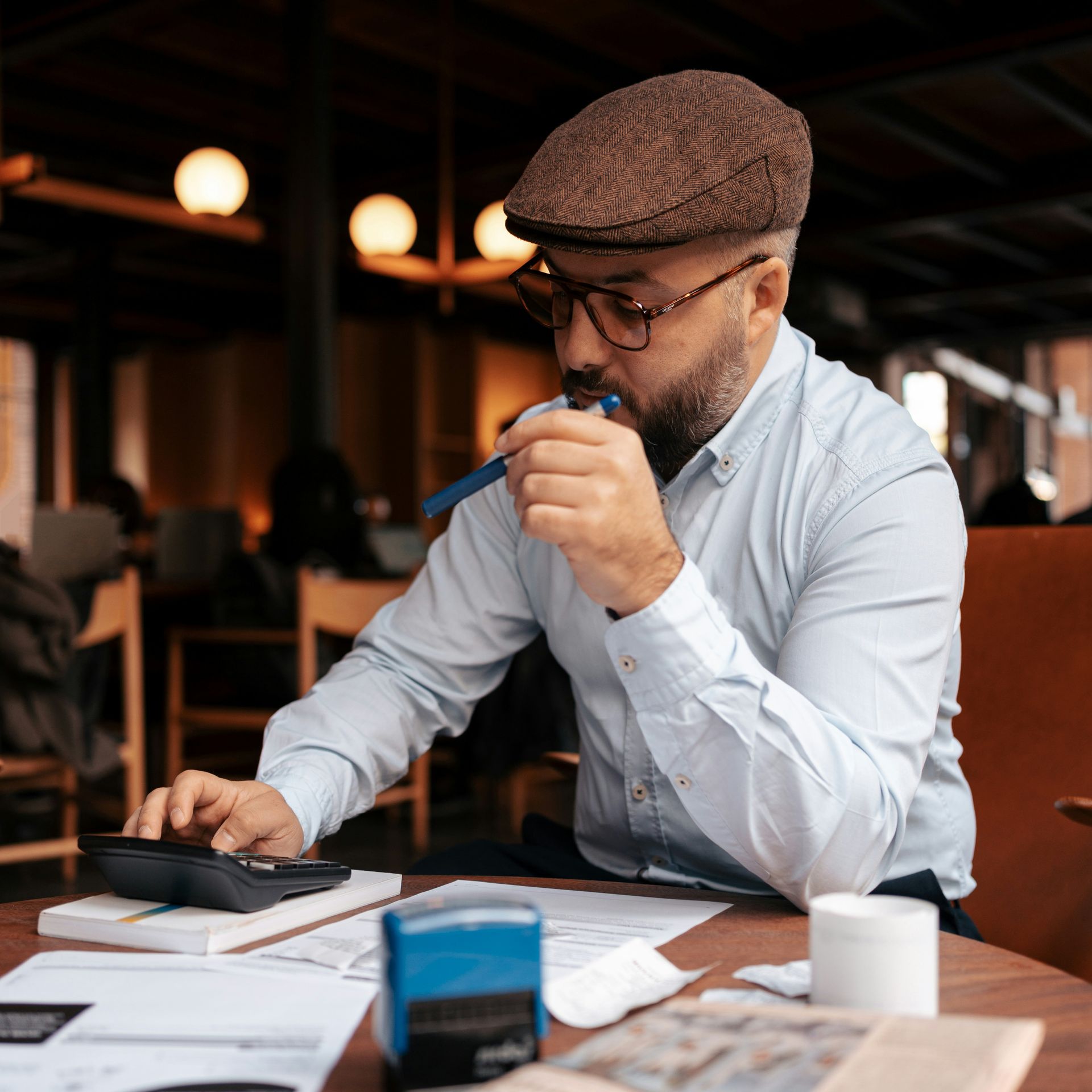 Man with glasses and cap calculating bills at a table in a cafe.