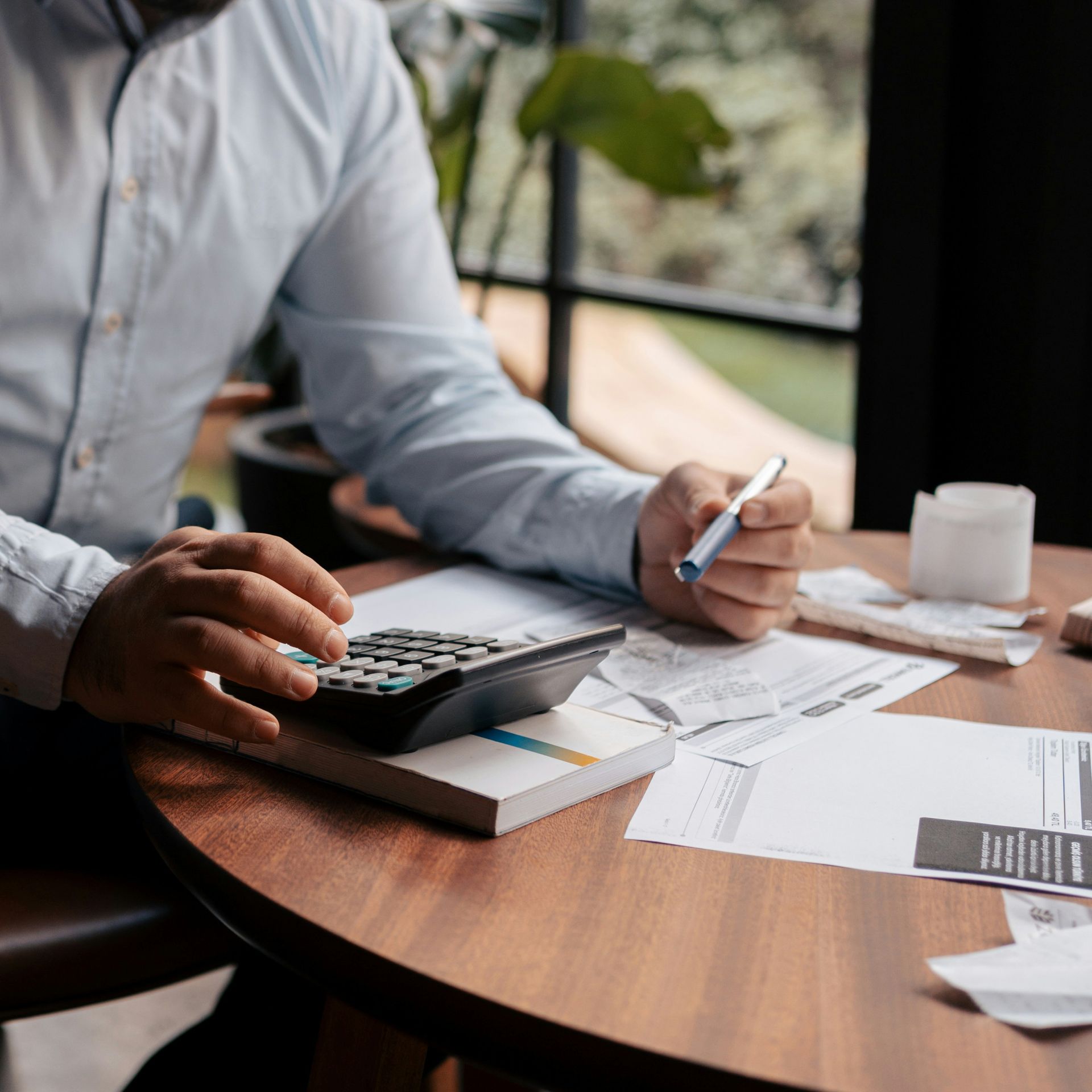 Man using calculator and pen at table with documents, calculating finances indoors.