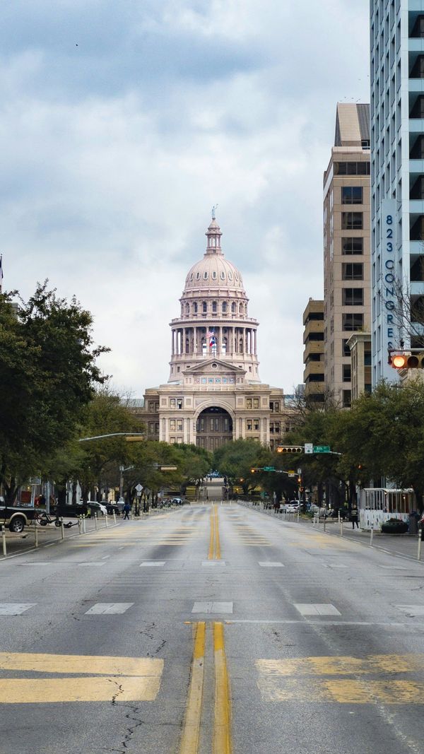 Texas State Capitol building at the end of a city street with trees on either side. Cloudy sky overhead.