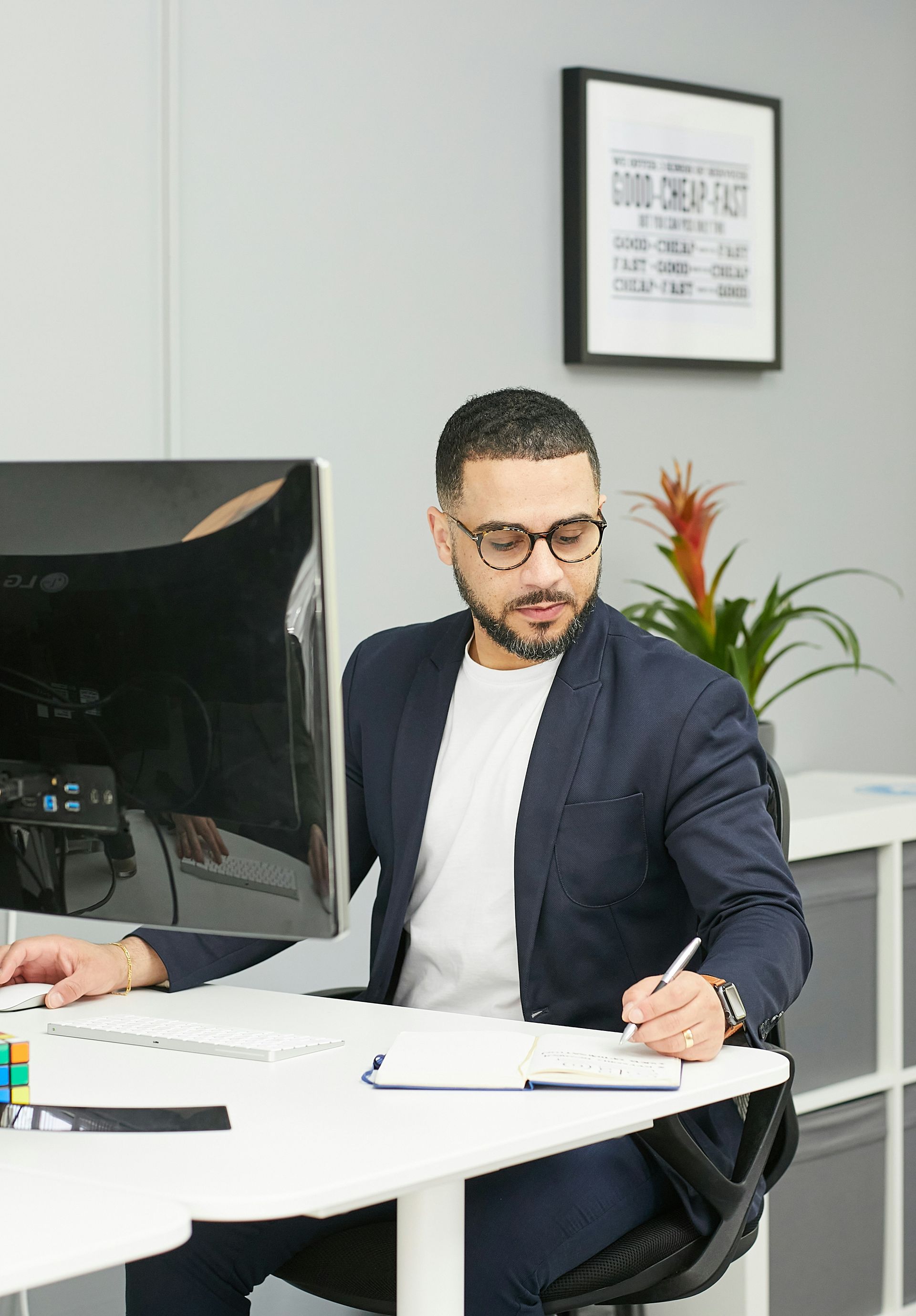 Man in glasses and blazer sitting at a desk, writing and looking at a computer monitor.