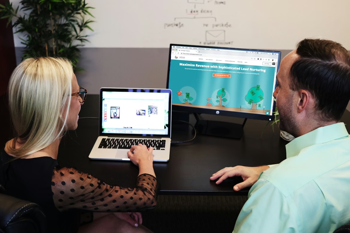 Two people reviewing a laptop and monitor with a website open in an office.