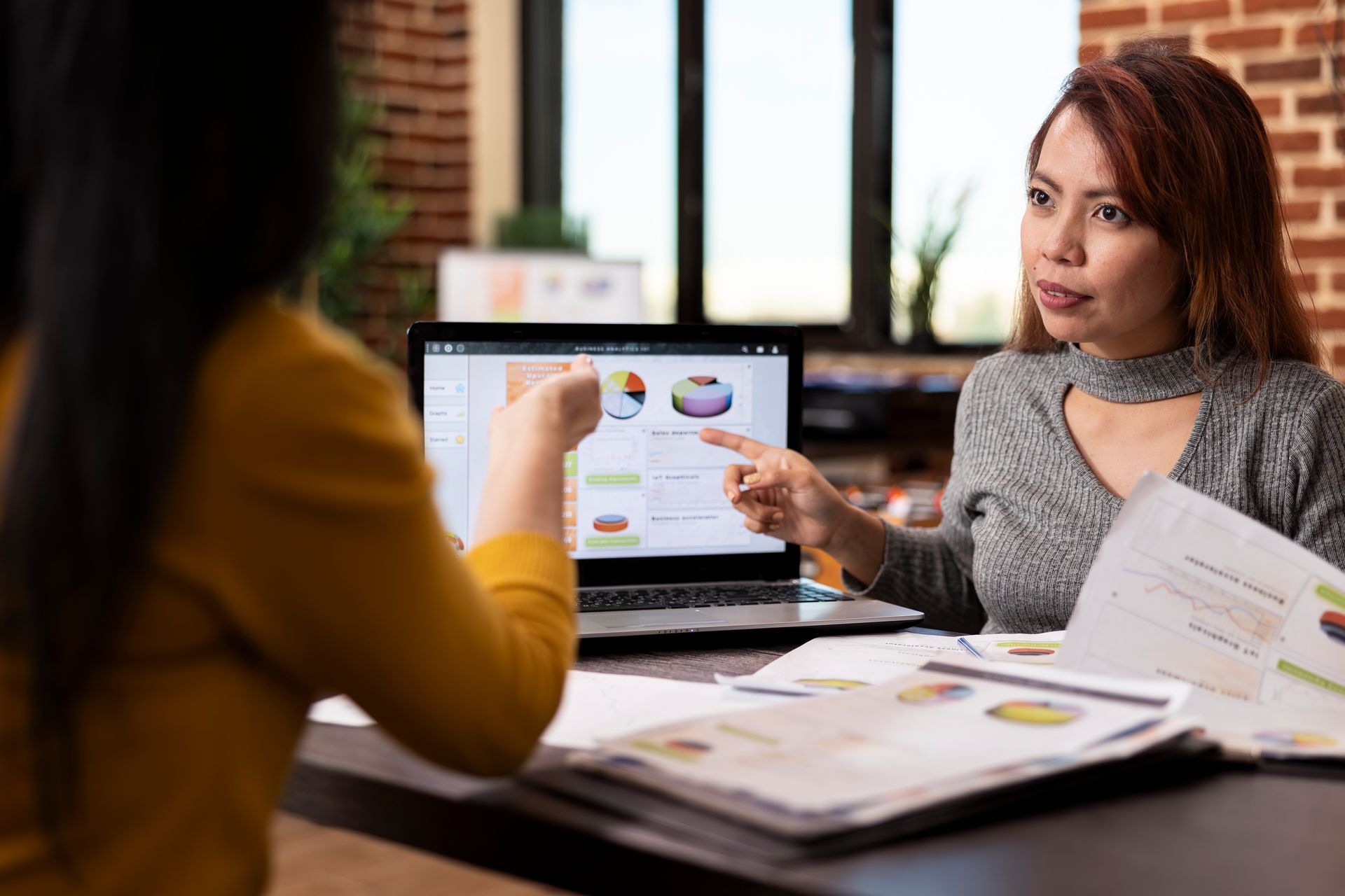 Two people discuss charts and documents at a desk in a bright office meeting room.