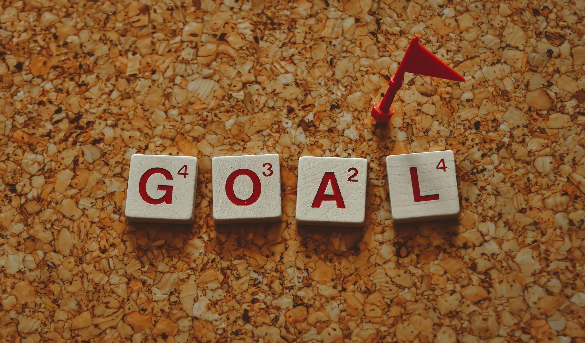 Scrabble tiles spelling “GOAL” with a red flag on a corkboard background