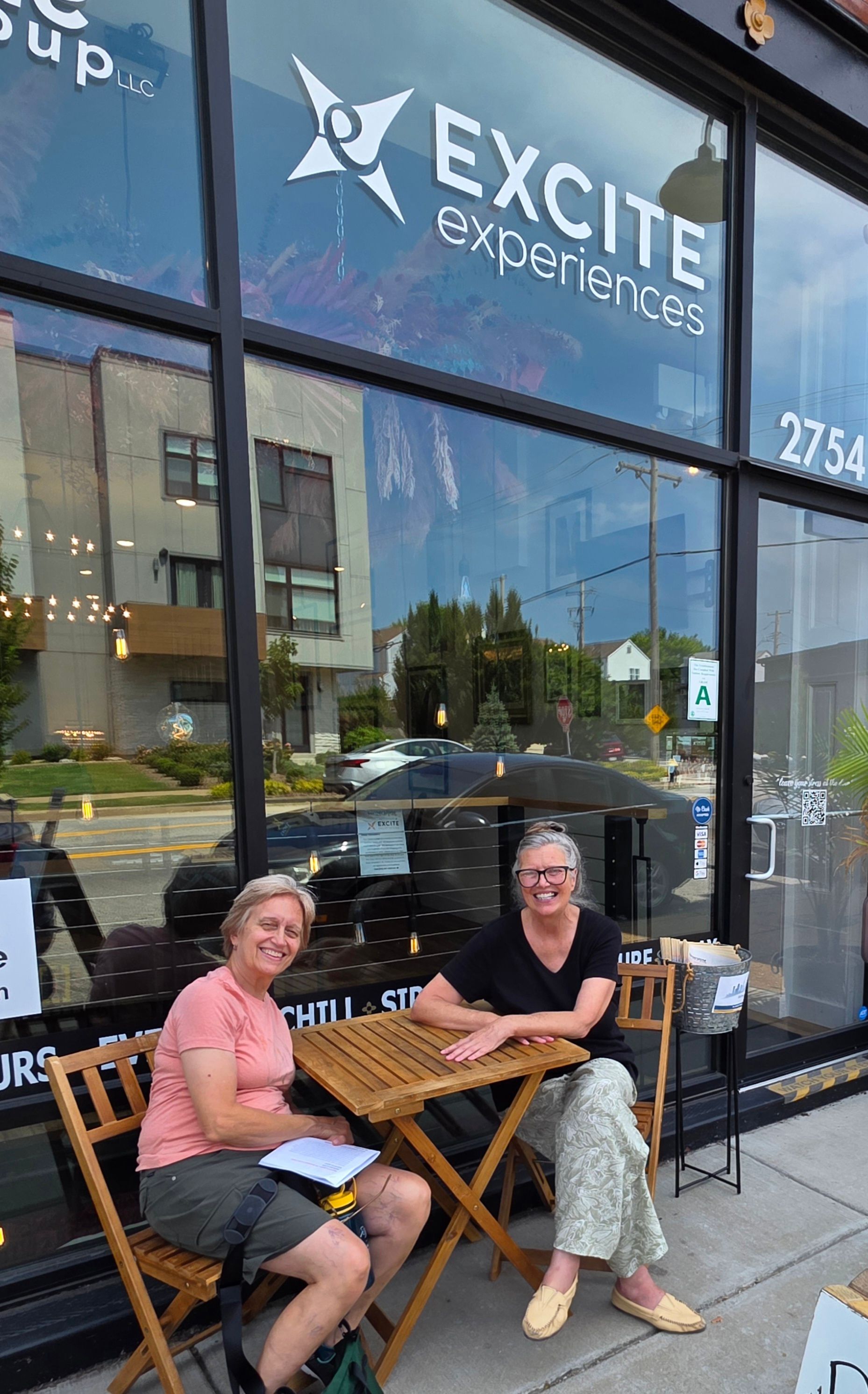 Two people sit at an outdoor table outside EXCITE, smiling in front of a storefront window.