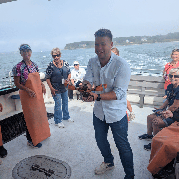 Man on boat holds up a lobster, smiling. Other tourists watch.