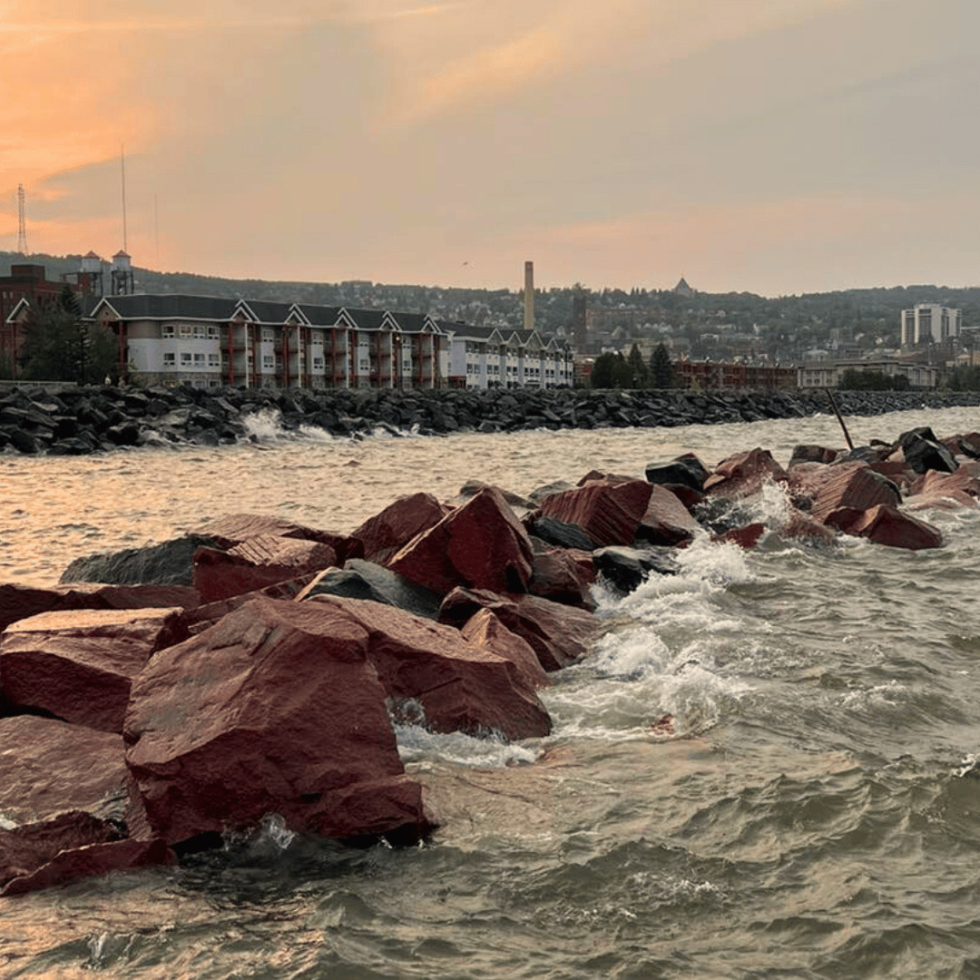 Waves crash on red rocks along a lakefront, with buildings and a skyline in the distance under a dusky sky.