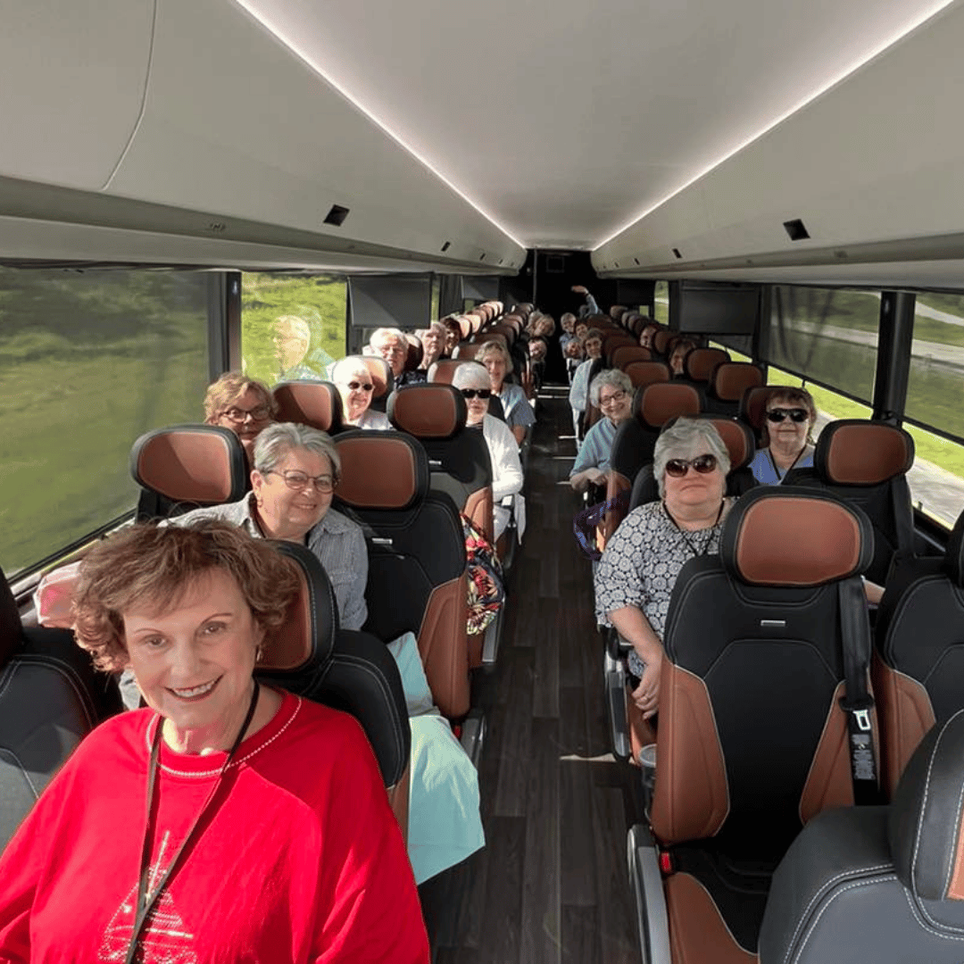 A bus interior filled with older adults, many wearing sunglasses. Woman in red smiling in the foreground.