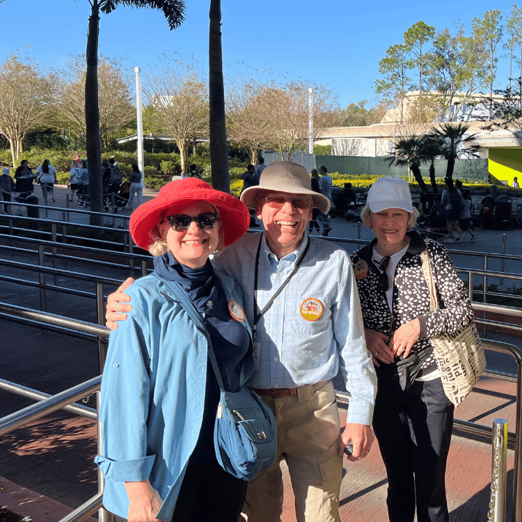 Three people smiling outdoors: woman in red hat and blue jacket, man in tan hat and light blue shirt, woman in white hat and patterned jacket.