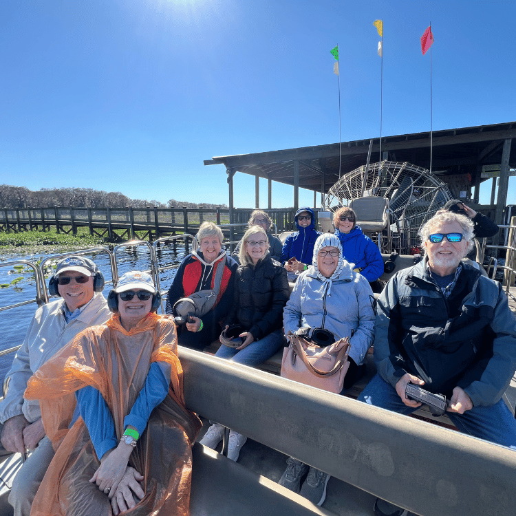 People ride an airboat on a sunny day. Passengers sit on benches; flags fly overhead.