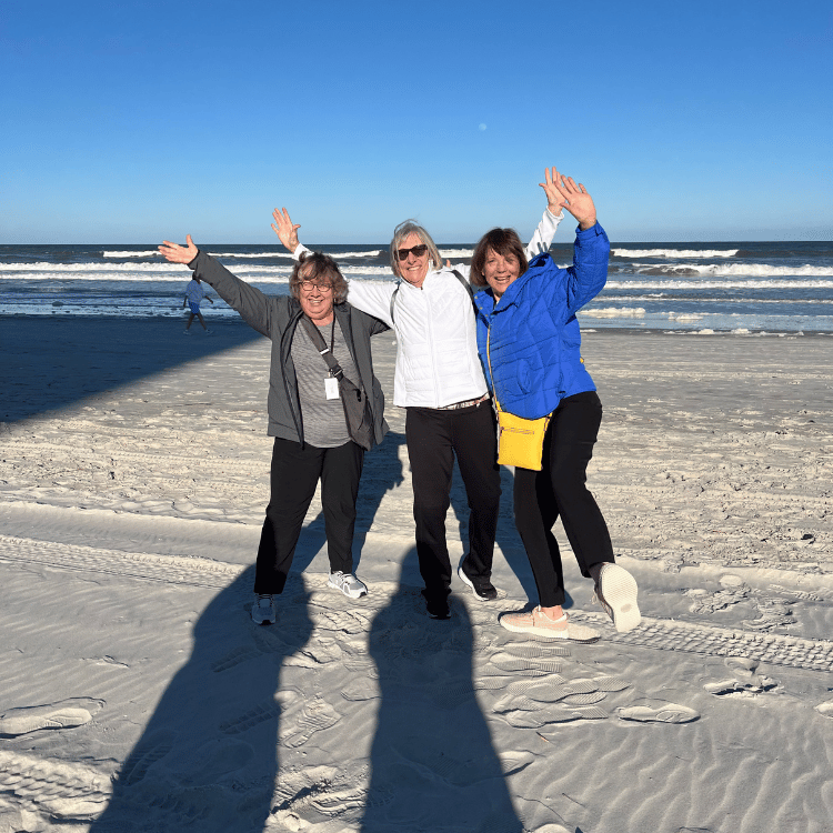 Three people on a beach with arms raised, sunny day.