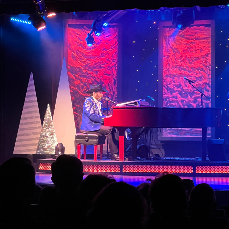 Man in blue jacket plays a red piano on stage, with audience in foreground.