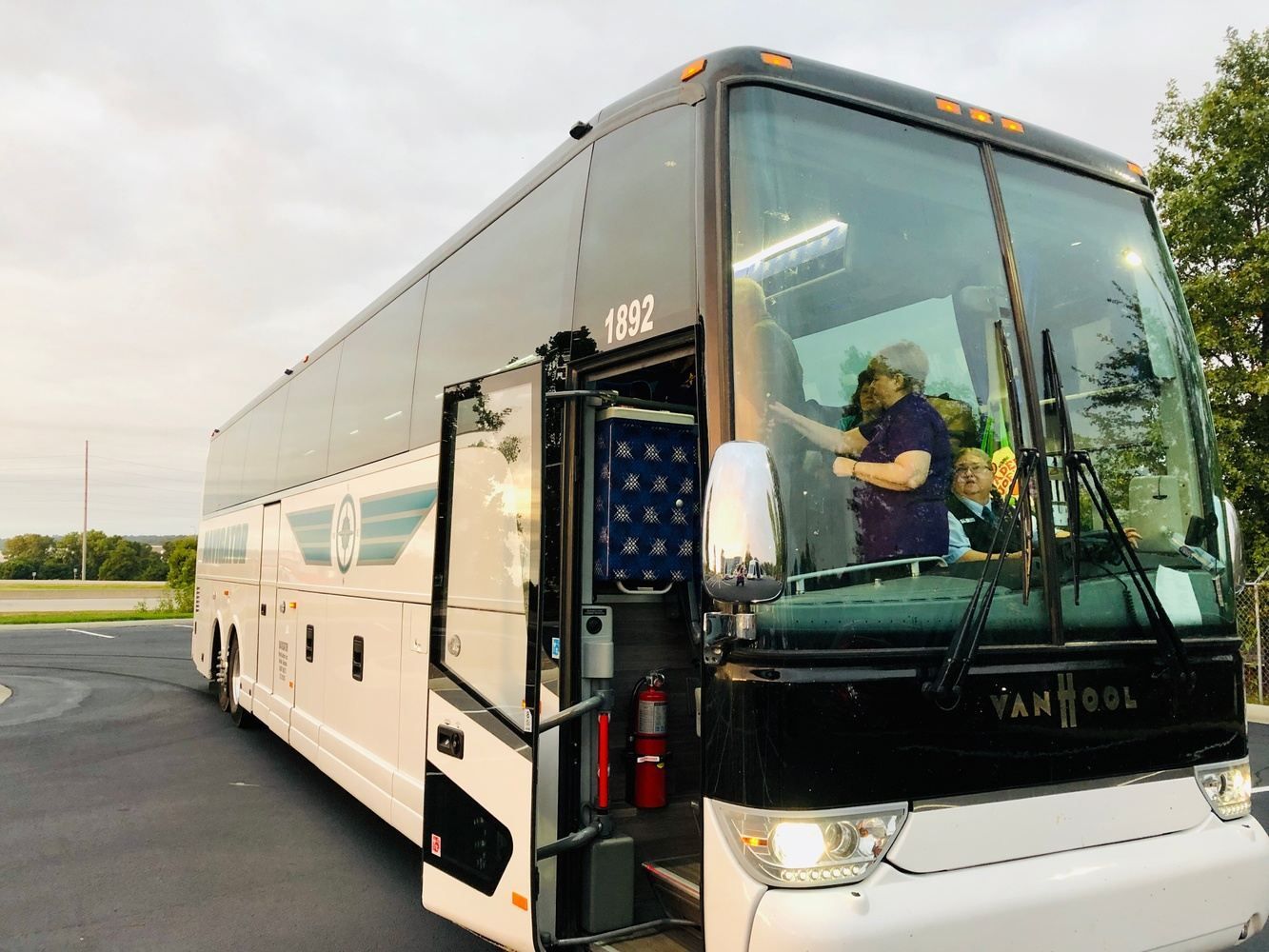 Gray and white tour bus with open door, driver and passengers visible. Evening setting.