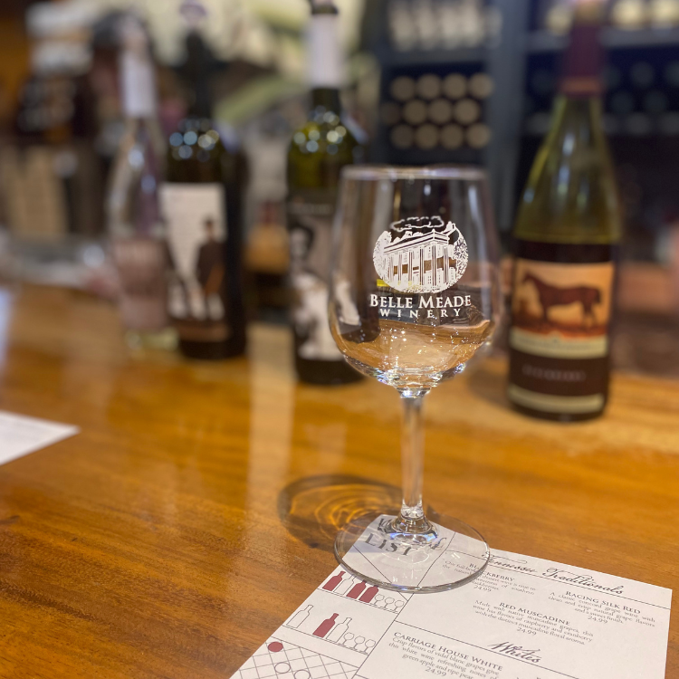 Wine glass with Belle Meade Winery logo on a wooden table, bottles in background.