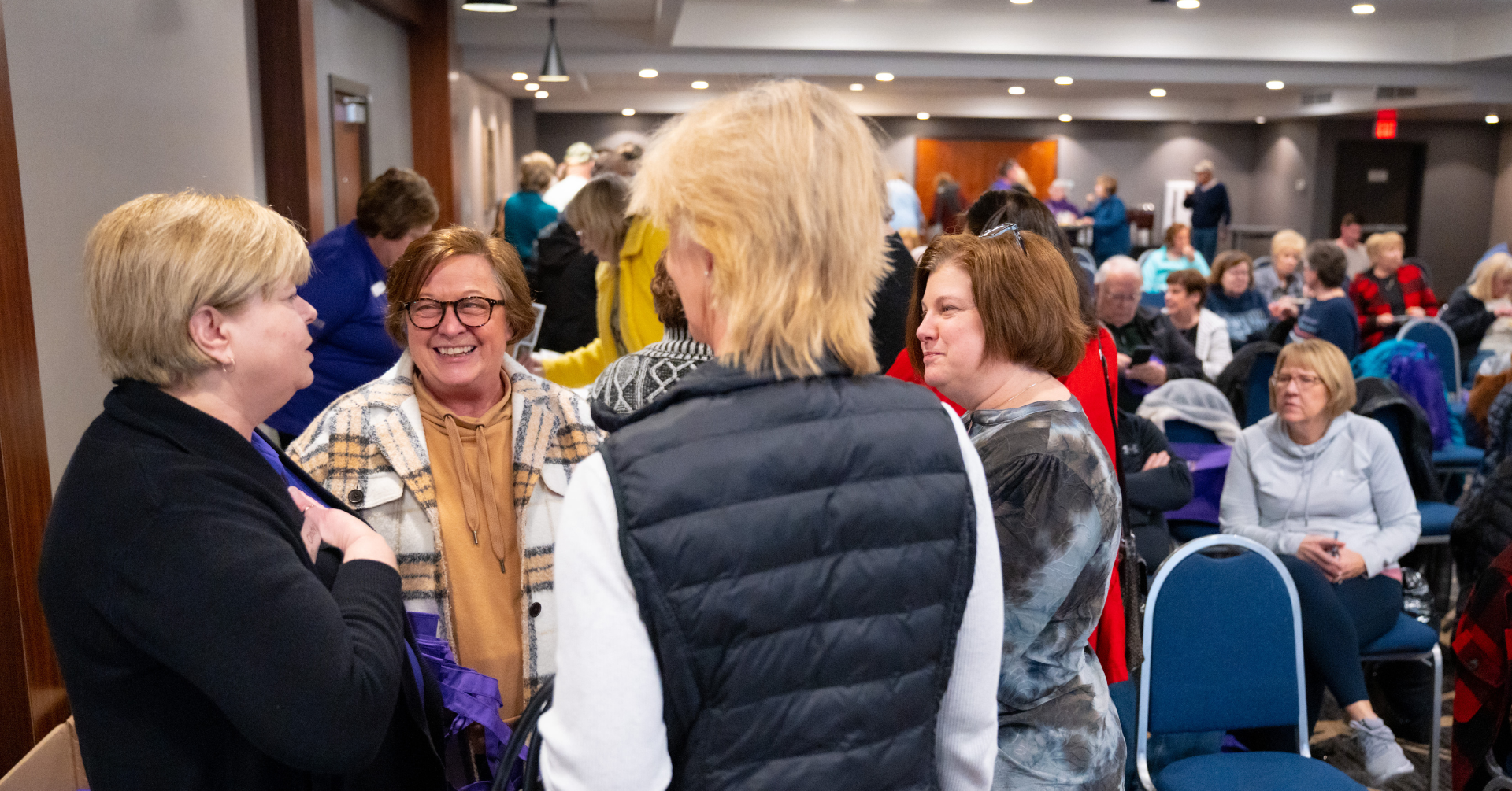 People in a room, socializing. Women in jackets and tops. Some seated. Neutral walls and blue chairs.