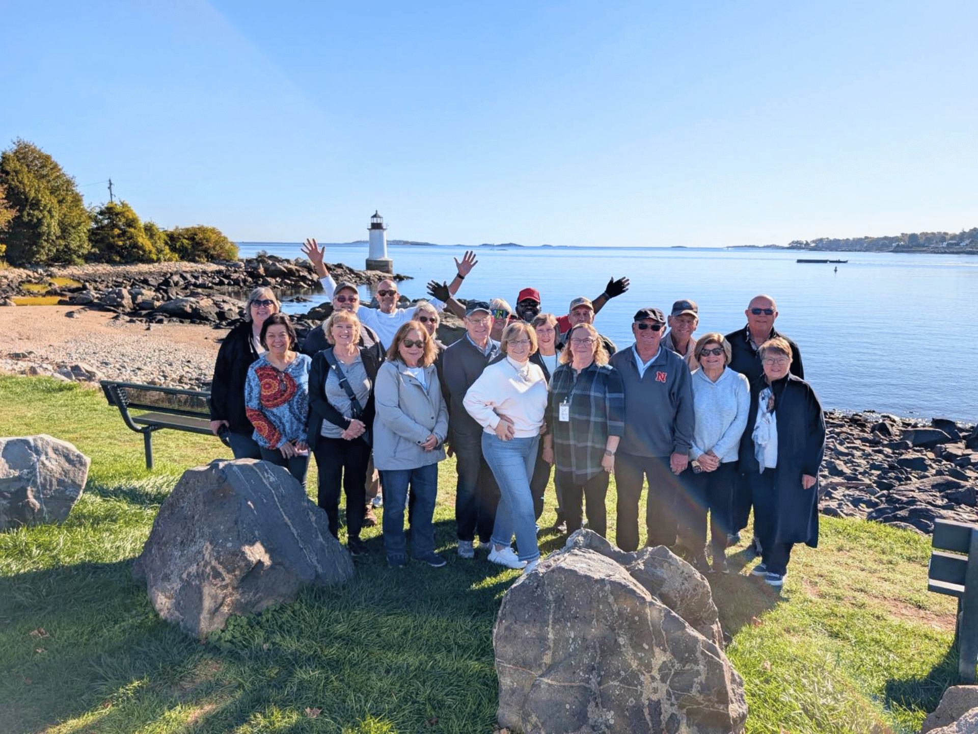 Group of people posing by a rocky shoreline and lighthouse on a sunny day.