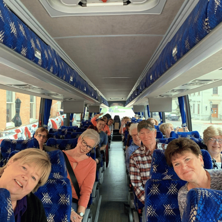 People on a bus, smiling. Inside with blue seats and overhead compartments, windows, outside view.