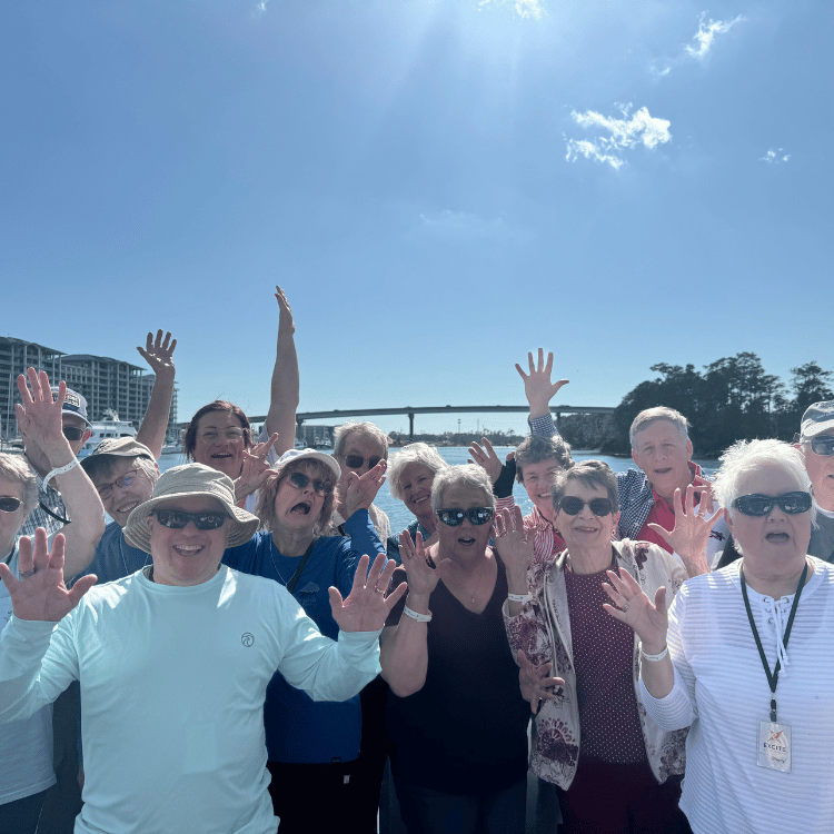 Group of people waving on a sunny day near water, bridge in background.
