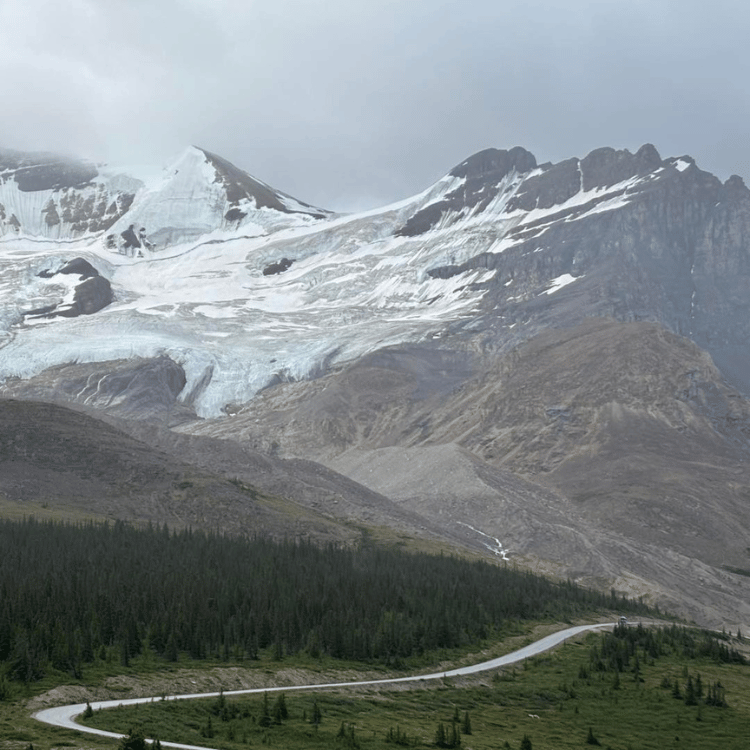 Mountain with a glacier under a cloudy sky; road winds through the valley.