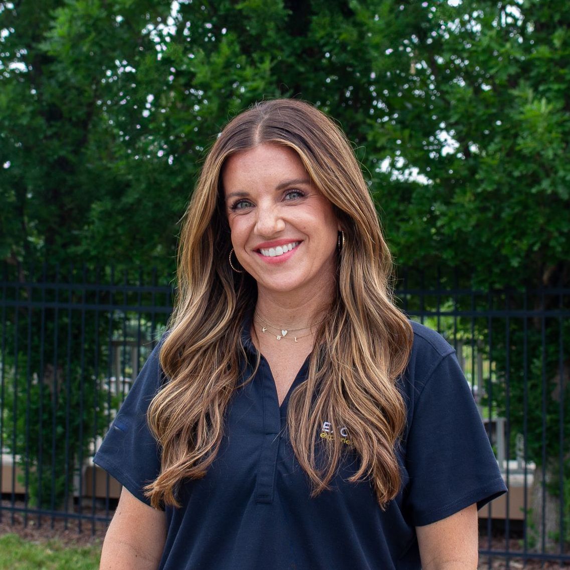 Woman with long brown hair smiling, wearing a dark blue top, in front of greenery.