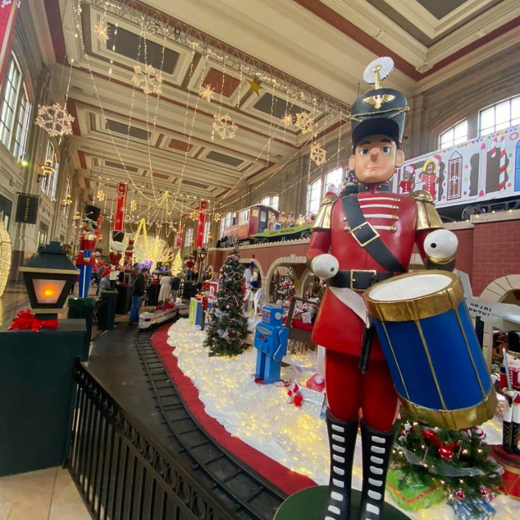 Toy soldier stands by train display, Christmas decorations in a grand hall.