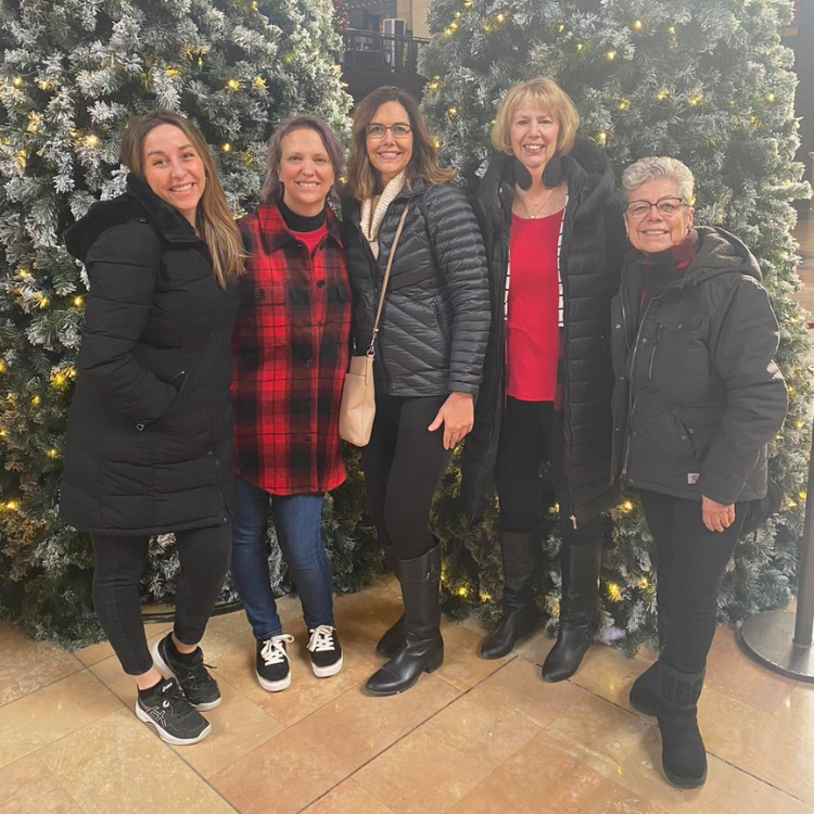 Five women pose by Christmas trees; smiling, dressed warmly, inside.