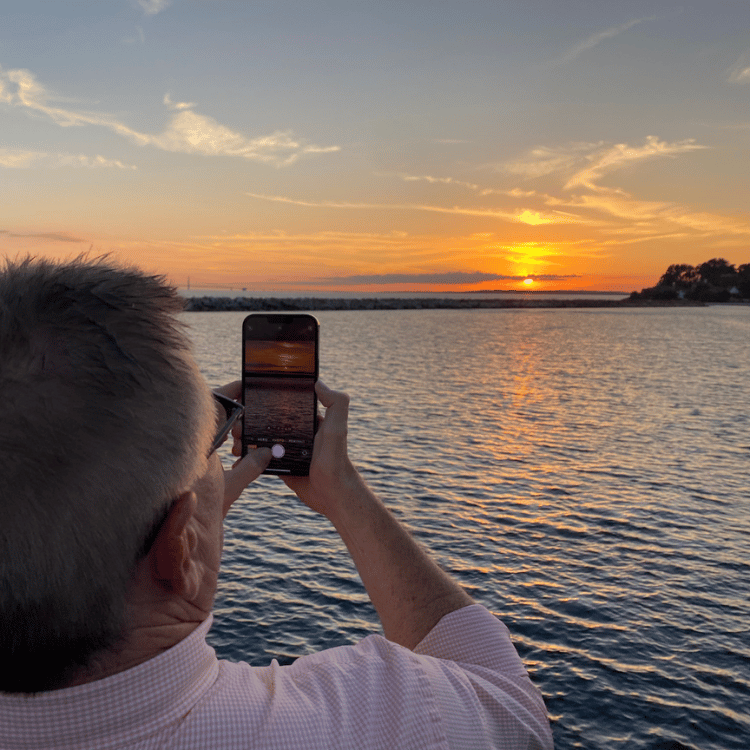 Man photographing a sunset over water with a smartphone. Orange sky, calm water.