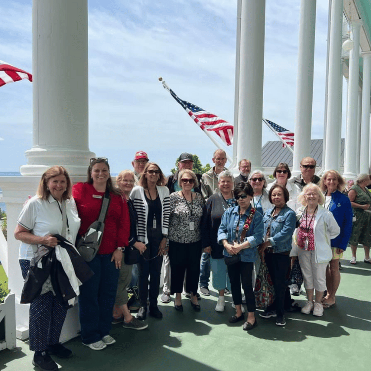 Group of tourists posing on a porch with American flags. Smiling people, white columns, and blue sky.