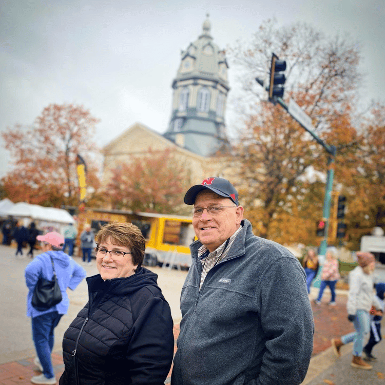 Couple posing on a street with a domed building in the background, fall trees and people.