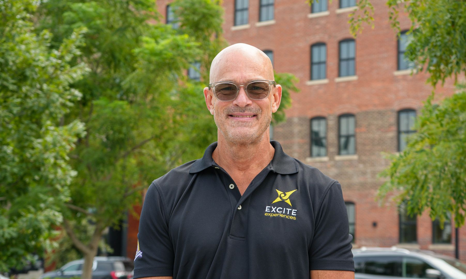 Man with glasses and shaved head smiles, wearing a black polo shirt, against a wood-grain background.