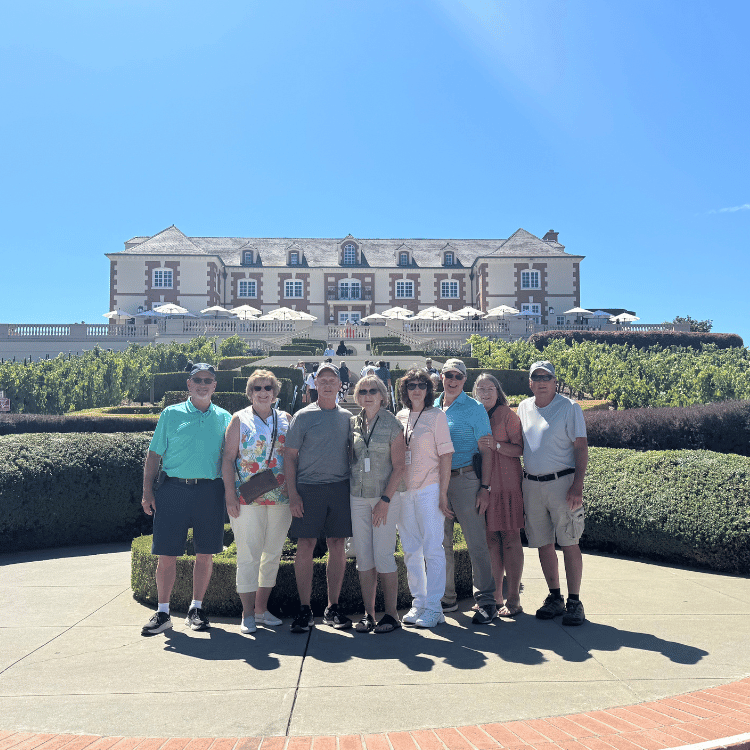 Group of nine people smiling in front of a large, white building with a vineyard on a sunny day.