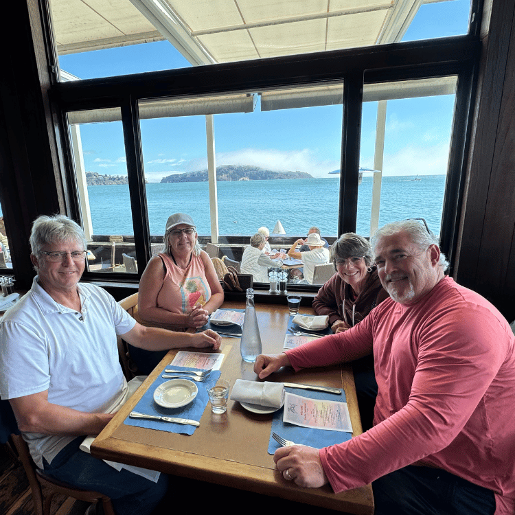 Four people at a table in a restaurant by a window with a view of the water and a partially clouded island.