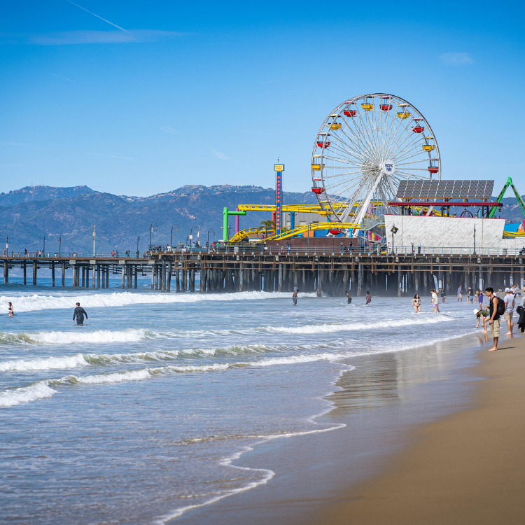 Beach scene with Santa Monica Pier, Ferris wheel, and mountains in the background under a blue sky.