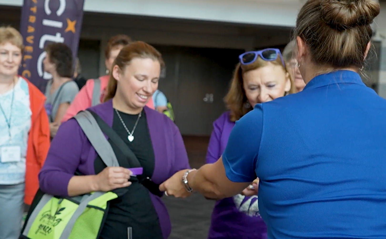People at an event checking in, woman in purple receives card from another woman in blue shirt, other people in background.