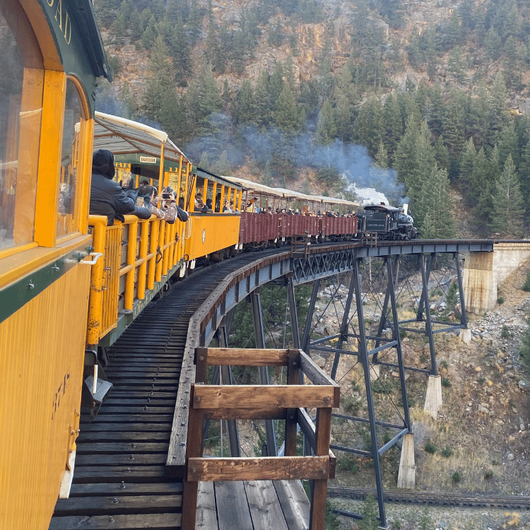 Train traveling over a high trestle bridge in a mountainous area; passengers aboard in yellow cars.
