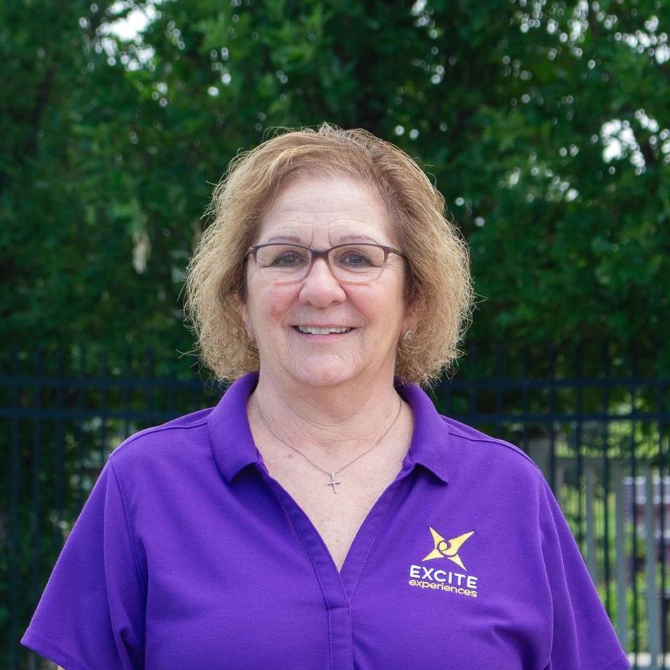 Woman in purple shirt with glasses, smiling in front of a fence and green foliage. 