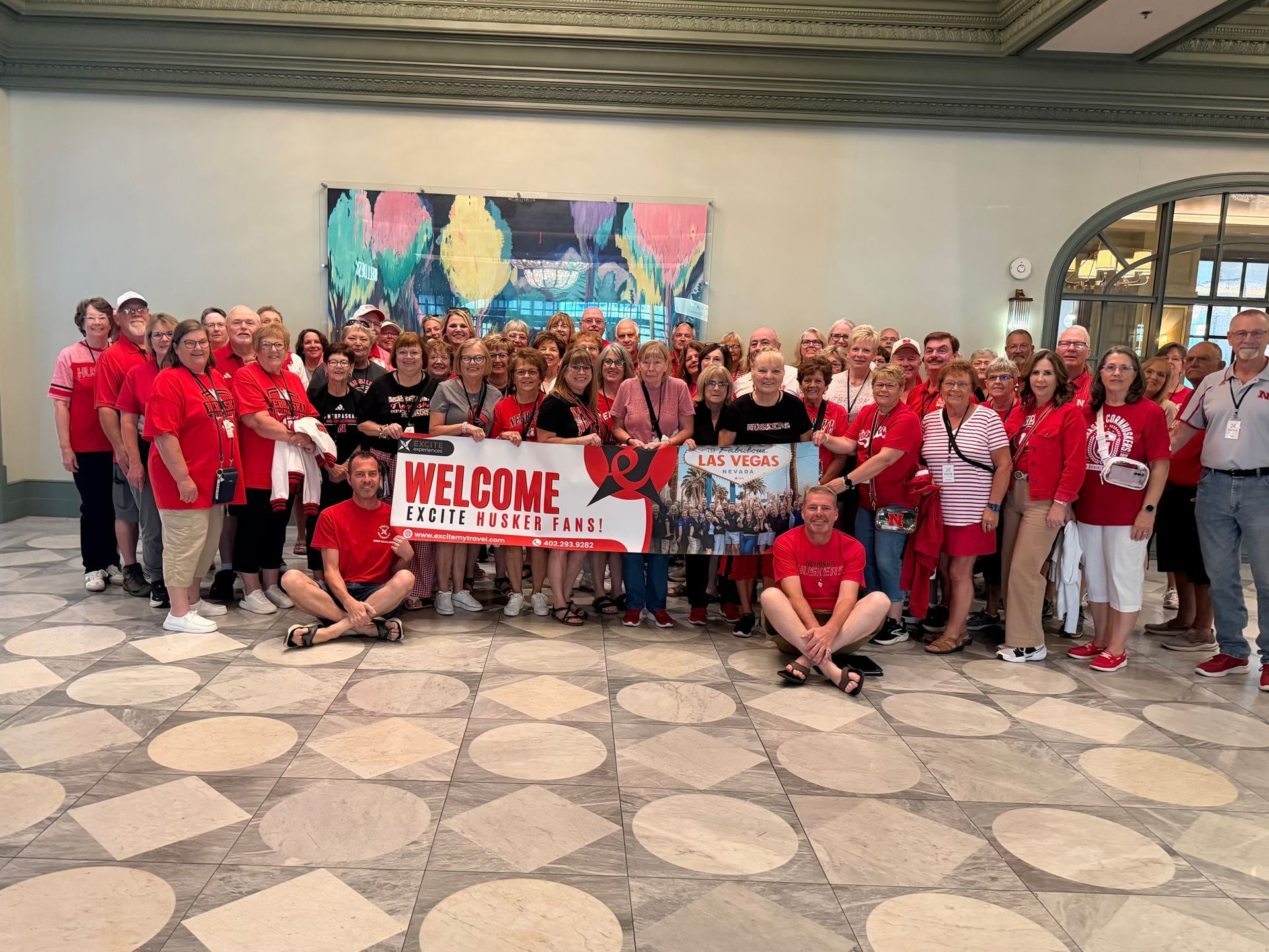 Large group in red shirts posing indoors with a “WELCOME” banner in front of a colorful mural